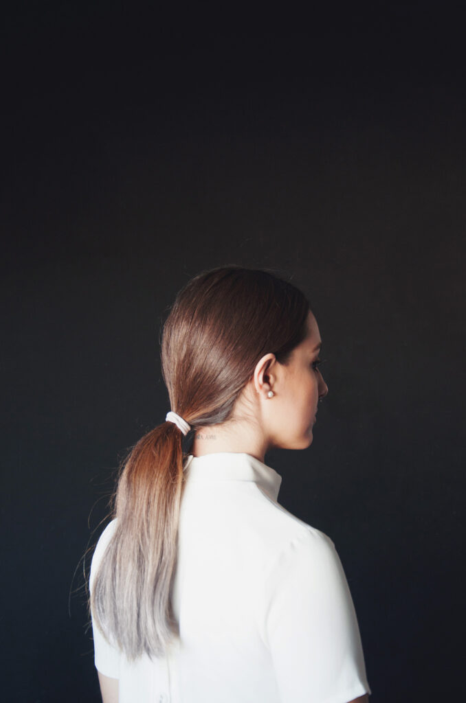 Laura Adriana photo profile view of a person with brown hair tied in a ponytail wearing a white shirt against a dark background.